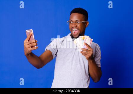 Young handsome African man on glasses taking selfie with his mobile phone and holding cash, on an isolated background Stock Photo