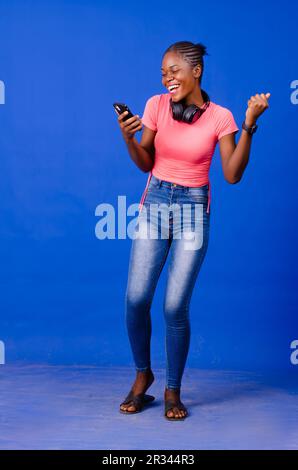 Young African lady dancing, listening to music and using mobile phone isolated on a blue background Stock Photo