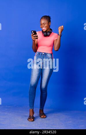 Happy Young African lady dancing, listening to music and looking at her mobile phone isolated on a blue background Stock Photo