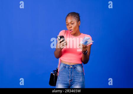 Young African lady holding cash and using mobile phone, isolated on a blue background Stock Photo