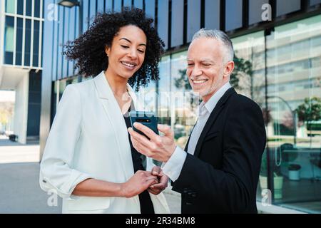 Two business people using a cellphone to watch the corporate shares. Businessman showing his smartphone app to his businesswoman partner. Corporate colleagues wearing formal suit talking at workplace. High quality photo Stock Photo