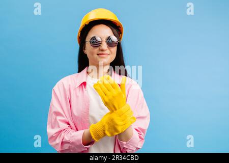 Woman in yellow helmet and gloves pointing and gesturing against blue studio background. Stock Photo