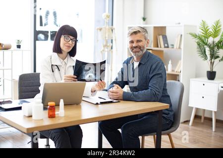 Portrait of happy doctor and patient sitting at table with portable computer in modern medical office and smiling at camera. Qualified radiologist dressed in white lab coat holding x-ray in hands. Stock Photo