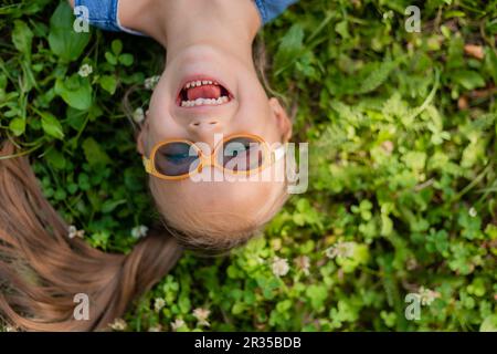 happy little girl lying on the green grass in summer top view. A child with down syndrome wearing glasses for vision. High quality photo Stock Photo