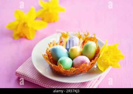 A caramel nest with colourful sugar eggs on a plate, and daffodils in the background Stock Photo