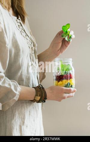 A woman in a linen dress holding a jar of fruit salad with mango, pomegranate seeds and lambs lettuce Stock Photo