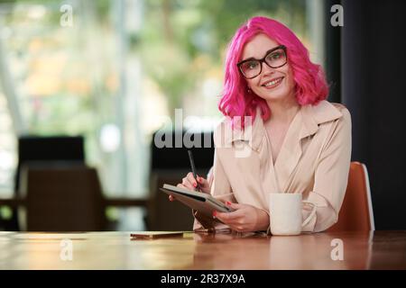 Portrait of happy young entrepreneur in glasses filling planner on tablet computer when drinking morning coffee Stock Photo