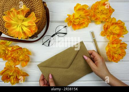 Women's hands are holding a craft envelope. On a white wooden table there is a wicker bag and yellow terry tulips.  Stock Photo