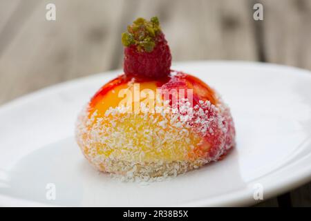 A mango and passion fruit mousse on a pastry crust with shredded coconut topped with a raspberry and chopped pistachio nuts on a white plate Stock Photo