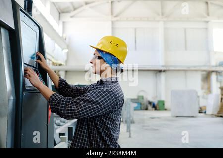 Female worker in hard hat pushing buttons on machine and looking at screen of computer while working in plant Stock Photo