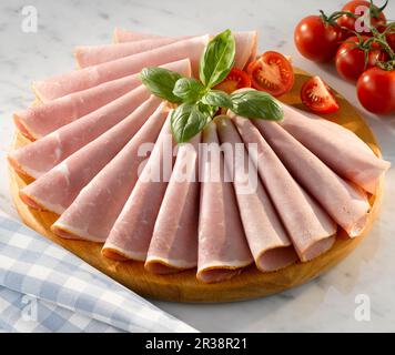 Slices of honey roasted ham on a circular wooden cutting board with garnish of basil and tomatoes Stock Photo