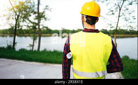 Construction worker wearing yellow vest staying in front of the lake. Worker holding construction plan. Stock Photo