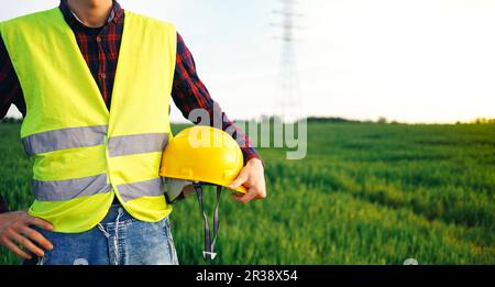 Construction worker holding yellow hard hat and wearing yellow fluorescent waistcoat. Electrician staying on the field. Construction site. Stock Photo