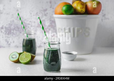 Spirulina and green fruit smoothie in glass jars, halved kiwi and limes, a big white bowl of assorted fruit on a light colored tabletop Stock Photo