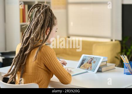 Rear view of student with dreadlocks making notes while sitting at table in front of tablet pc and having video call with teacher Stock Photo