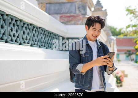 Portrait of Asian male traveler using a mobile phone on sidewalk of buddhist temple on street in Bangkok, Thailand, Southeast Asia - smart phone and i Stock Photo