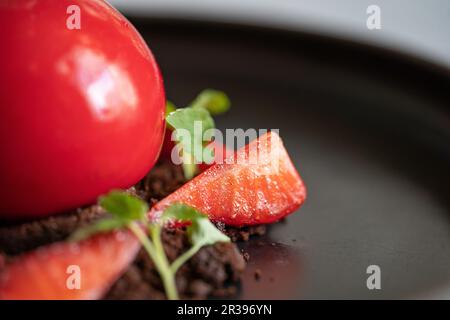 Strawberry dessert detail. Sphere shaped curd cake with strawberries and brownie crumbs. Red dessert on the black plate. Selective focus. Stock Photo
