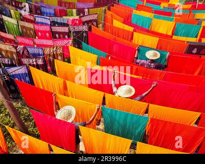 Narayanganj, Dhaka, Bangladesh. May 23, 2023, Narayanganj, Dhaka, Bangladesh: Workers hang hundreds of color-dyed sheets of cloth on a bamboo framework to dry in a dyeing factory in Narayanganj, Bangladesh. The drying process usually takes 4 hours, with each set of 200 pieces at a time to dry in temperatures over 42 degrees Celsius. Workers use hats for protection from the scorching heat because they have to constantly turn the colorful fabrics so that they dry perfectly in the sunlight. Credit: ZUMA Press, Inc./Alamy Live News Stock Photo