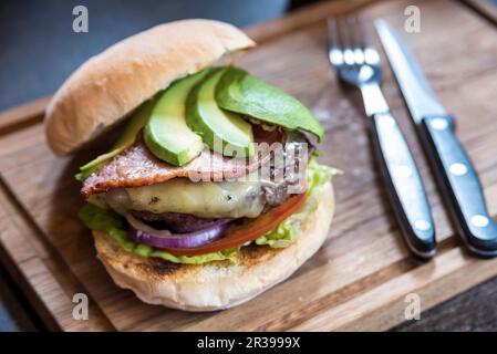 Burger with avocado slices, cheese, bacon, onions, lettuce, tomatoes on a wooden board with cutlery Stock Photo