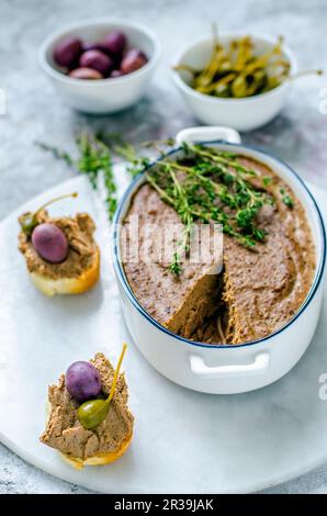 Liver pate in ceramic form with fresh thyme, and two baguette sandwiches with pate, capers and olives on a marble stand Stock Photo