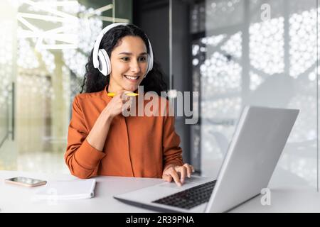 Young curvy businesswoman with headphones listening to online course and audio podcast female worker inside office working with laptop, successful hispanic woman smiling satisfied with work. Stock Photo