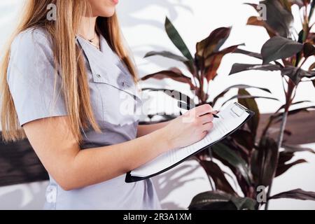 Closeup of female doctor in gray uniform writing down her patient details on paper form in modern clinic. Young beautician makes checklist for her cli Stock Photo