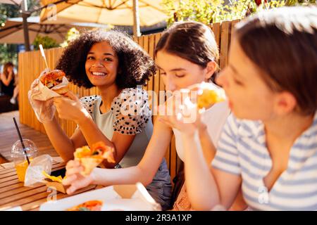 Cute smiling teenage girls are sitting in open air cafe and eating fast food. Cheerful happy female friends of different nationalities snacking on bur Stock Photo