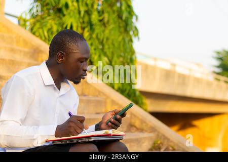 Business man seated writing on a notepad and using his mobile phone Stock Photo