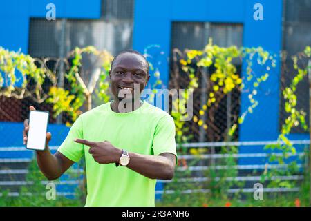 copy space image of excited african man wearing green t-shirt holding and pointing to a mobile phone with white blank screen. empty space for advert p Stock Photo