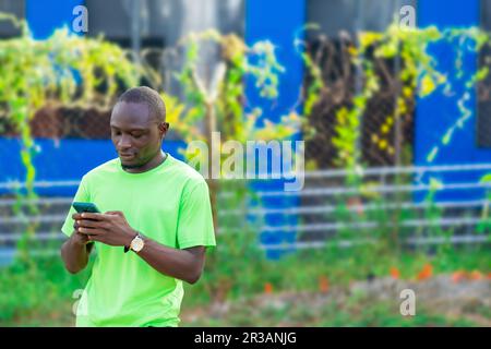 young african adult wearing green shirt standing the street alone and engrossed in a game on his technology device Stock Photo