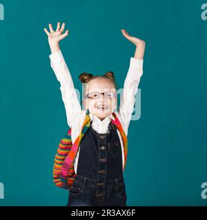 Funny smiling little girl with big backpack jumping and having fun against blue wall. Looking at cam Stock Photo