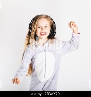 Cute little girl in headphones is using a smartphone, looking at camera and having fun at home. Day Stock Photo