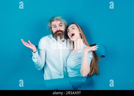 Surprised couple of bearded man and happy woman making hole in paper looking through hole. Advertising board, blue background. Couple through hole in Stock Photo