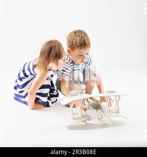 Little Children playing with paper toy airplane sitting on the floor against a white background. Sma Stock Photo