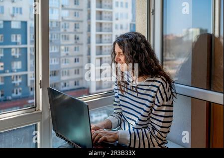 Smiling woman using laptop sitting near window Stock Photo