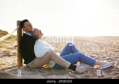 Smiling mature couple spending leisure time sitting on sand at beach Stock Photo