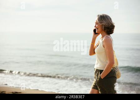 Smiling woman with hand in pocket talking on smart phone at beach Stock Photo