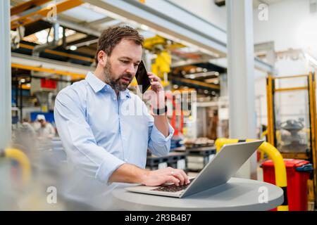 Manager talking on mobile phone using laptop at factory Stock Photo