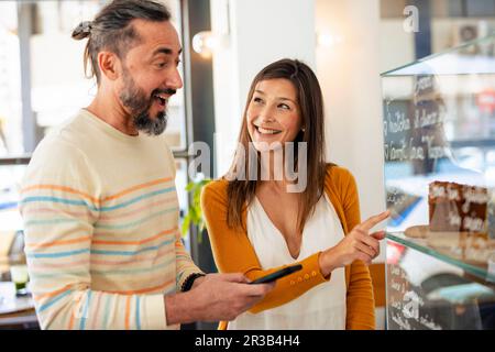 Smiling woman pointing at food display by man in coffee shop Stock Photo