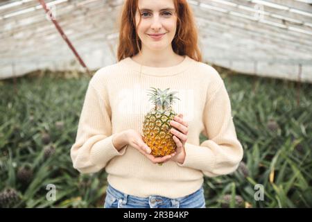 Smiling redhead woman holding pineapple in greenhouse Stock Photo