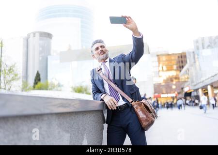 Businessman taking selfie through smart phone leaning on wall Stock Photo