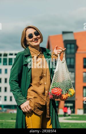 Stylish woman standing with vegetables in mesh bag Stock Photo