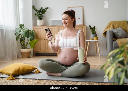 Smiling pregnant woman using smart phone sitting on exercise mat at home Stock Photo