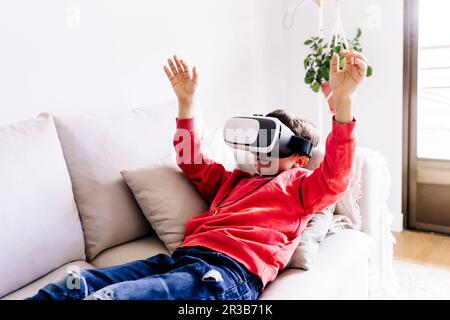 Boy with arms raised wearing virtual reality headset lying on sofa at home Stock Photo