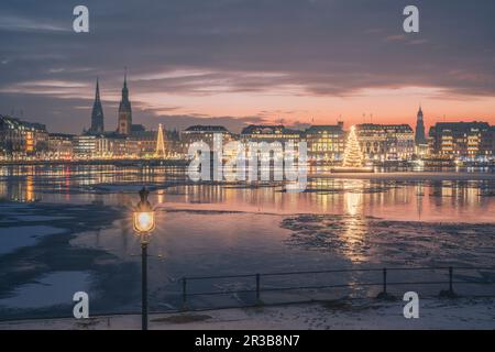 Germany, Hamburg, Ice floating in Alster Lake at dusk with city skyline and glowing Christmas trees in background