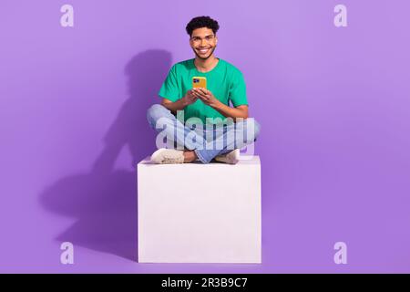 Full body photo of youth man enjoy his modern samsung smartphone four digital cameras sitting podium isolated on violet color background Stock Photo
