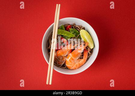 Asian stir fry soba noodles with shrimps, vegetables, green peas, red pepper in white bowl with wooden chopsticks Stock Photo