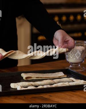 Woman twisting cinnamon bun dough into twists, on a baking sheet, with an oven in the background Stock Photo
