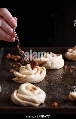 Cinnamon buns on a baking sheet, with a spoonful of cinnamon and brown sugar being placed on top Stock Photo