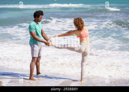 Man assisting girfriend standing on one leg at seashore Stock Photo
